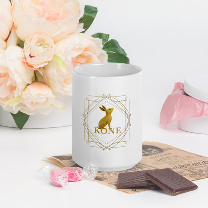 Taza KÖNE blanca con logo dorado ambientada en mesa de desayuno con flores y dulces.