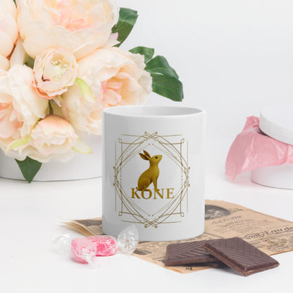 Taza KÖNE blanca con logo dorado en centro, colocada junto a flores rosadas y tableta de chocolate.